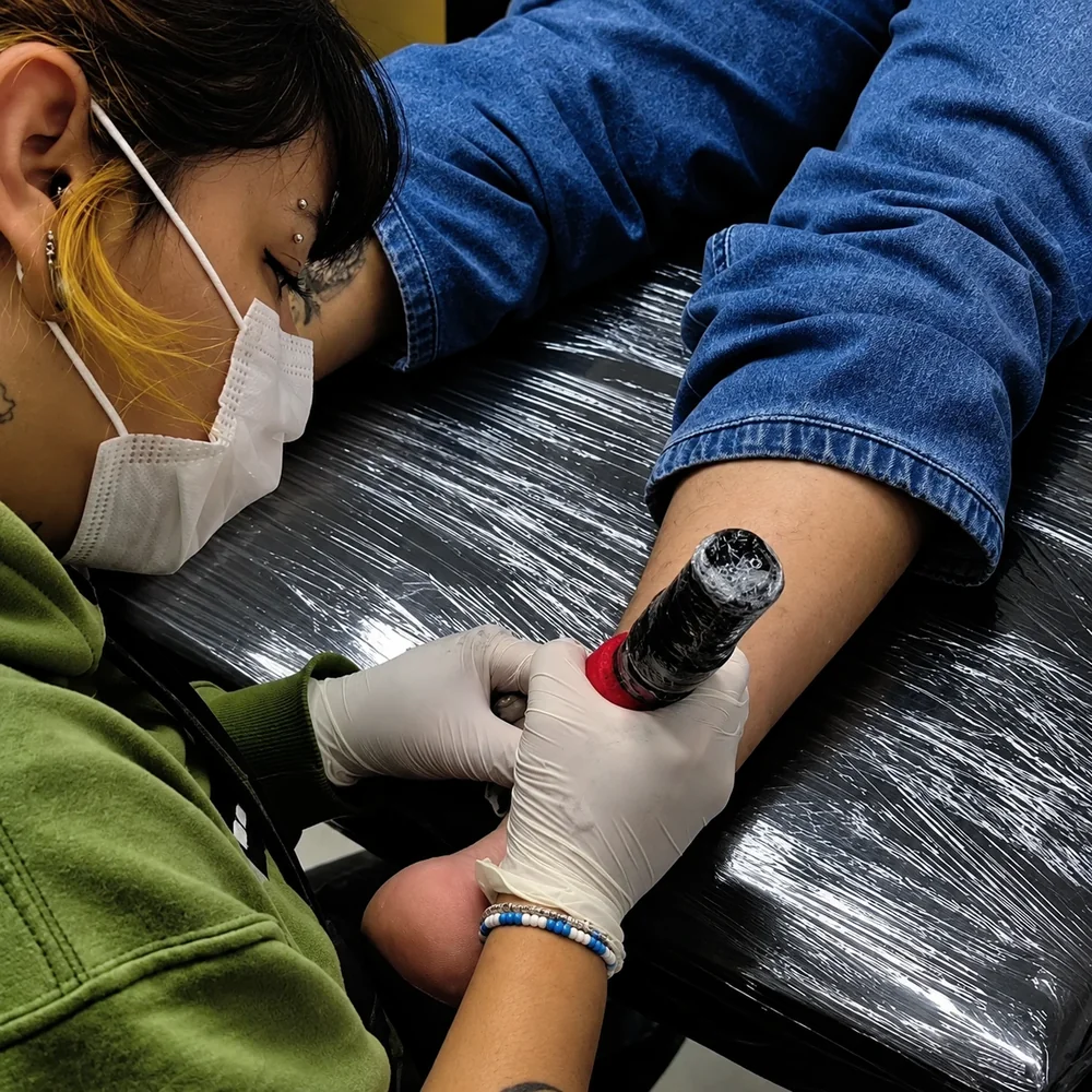 Diva tatuando um antebraço, de máscara e luvas